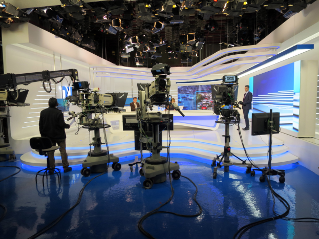 Ein TV-Studio mit mehreren Kameras auf dem Boden, einer sitzenden Person im Vordergrund und einigen stehenden Personen im Hintergrund, mit Deckenleuchten für eine Livesendung.