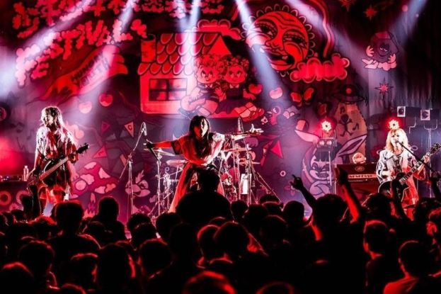 Ein Live-Auftritt einer japanischen Rockband mit Musikern, die Gitarren spielen und singen, auf der Bühne, begleitet von Mikrofonen und Ständern, während das Publikum unten zuschaut.