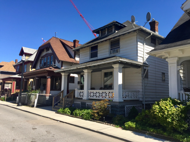 Eine Reihe von Häusern mit architektonischen Details wie Fenstern, Säulen, Geländern, Treppen, Dächern und einem Baukran auf einem Dach, neben einem Straßenmast, Pflanzen, einem Zaun, einem Weg, Bäumen und einem bewölkten Himmel.