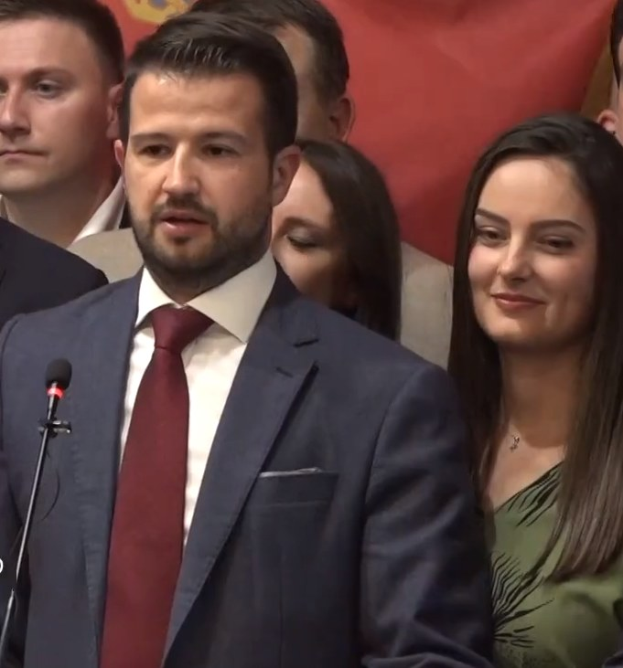 Ein Mann im Anzug und Krawatte steht an einem Podium mit einem Mikrofon und blickt auf eine Menge, während im Hintergrund ein Banner zu sehen ist.