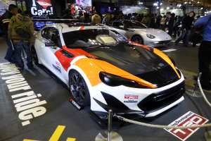 Eine Gruppe von Menschen betrachtet ein Rennfahrzeug auf der Tokyo Motor Show, umgeben von Absperrpollern mit Seilen und Text/Bild-Schildern im Hintergrund unter beleuchteter Decke.