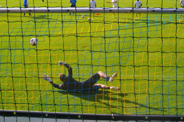 Fußballtorhüter hält vor einem Tor mit Netz im Vordergrund, einige Menschen stehen im Hintergrund und verstreute Fußballschuhe auf dem Boden.