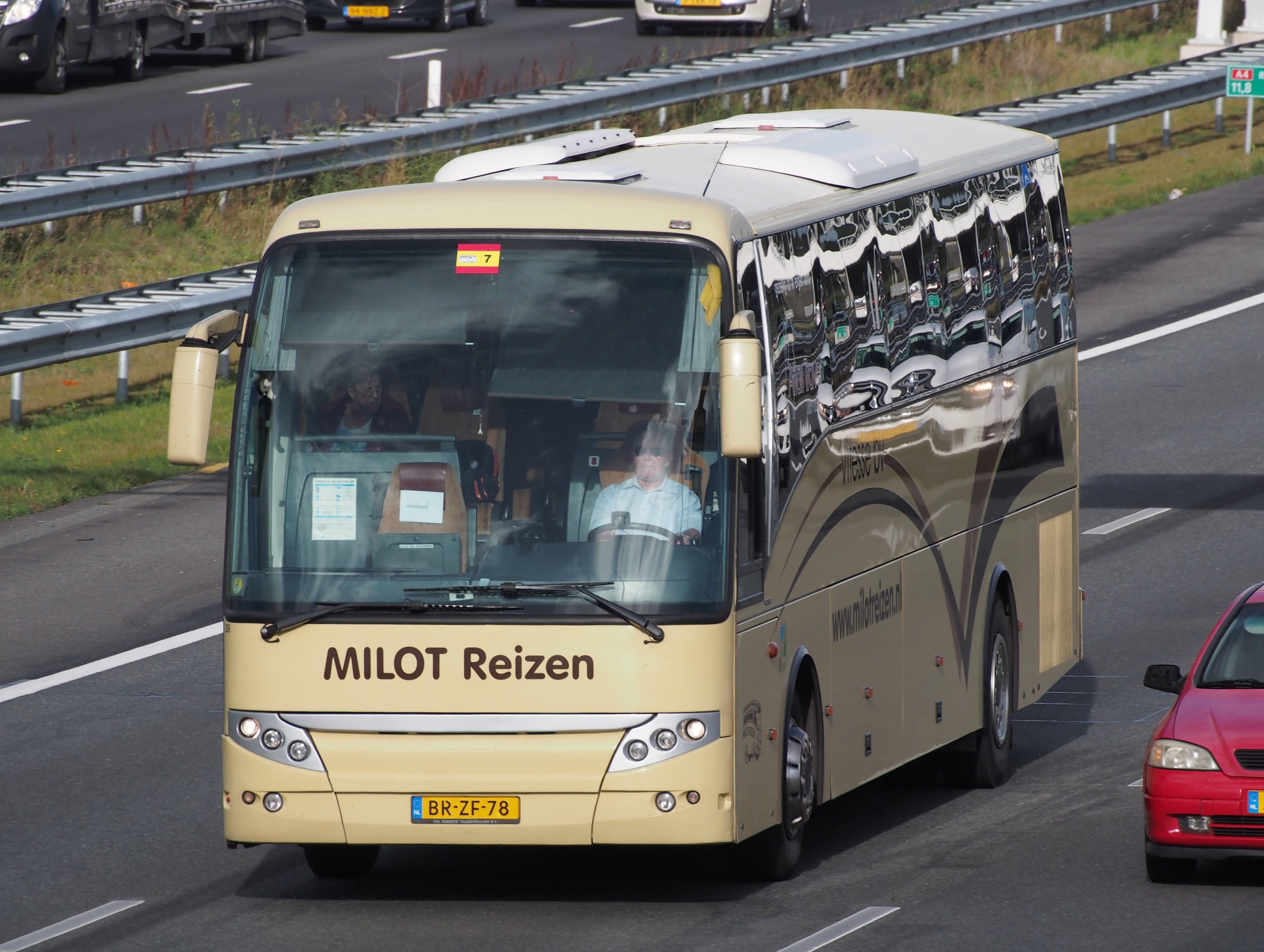 Ein Minibus mit Passagieren auf einer Autobahn neben einem roten Auto, mit Leitplanken und Gras an den Seiten und Säulen mit Text im Hintergrund.