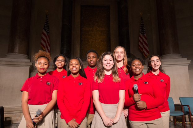 Gruppe junger Frauen in roten Hemden mit Mikrofonen, die lächeln, vor Stühlen, Säulen, Fahnen und einer Wand.