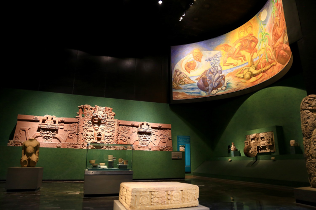 Ein Museumsraum mit verschiedenen Kunstwerken, darunter Skulpturen, Tischobjekte, eine Texttafel, eine Wandmalerei und Deckenbeleuchtung im Mexiko-Stadt-Museum für Anthropologie.