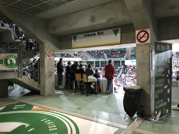 Gruppe von Menschen vor sitzendem Publikum in einem Stadion mit Schildern, die "Boston Celtics vs New York Jets" lesen, ein Müllcontainer, eine Tafel mit Text, eine Treppe mit Geländern und eine Decke mit Deckenleuchten.