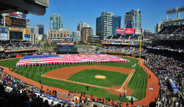 Baseballstadion voller Zuschauer, mit einer Gruppe unter einer amerikanischen Flagge auf dem Feld, Anzeigetafeln auf beiden Seiten und einer Kulisse aus Gebäuden, Bäumen und einem klaren Himmel.