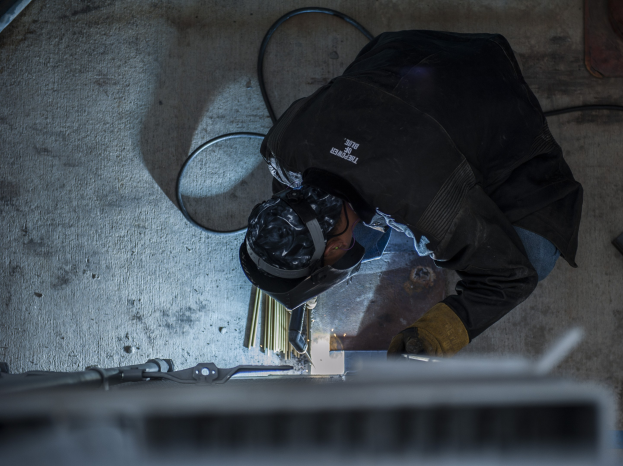 Ein Schweißer in Schutzausrüstung verwendet eine Schweißmaschine an einem Metallstück, mit Werkzeugen und Gegenständen, die auf dem Boden um sie herum verstreut sind.