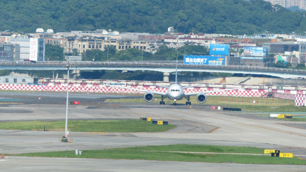 Flugzeug startet von einer Flughafenrollbahn mit umgebenen Infrastruktur wie Gras, Pfählen, Schildern, einem Zaun, Brücke, Wasser, Gebäuden und Bäumen unter einem klaren blauen Himmel.