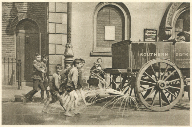 Schwarz-weißes Bild von Kindern, die vor einem Gebäude mit Fenstern, einer Tür, einem Mast und einem Zaun mit einem Feuerhydranten spielen, mit einem Wagen im Hintergrund.