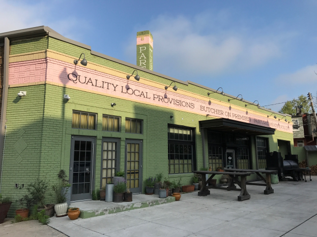 Außenansicht eines grünen Gebäudes mit einem "Qualität lokale Versorgung"-Schild, Fenstern, Türen, Topfpflanzen, Bäumen, Strommasten, Drähten und einem bewölkten Himmel.