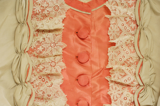 Nahaufnahme eines rosafarbenen und cremefarbenen Kleides mit weißer Spitzenverzierung und vorderen Knöpfen, das eine klassische und elegante Gestaltung aufweist.