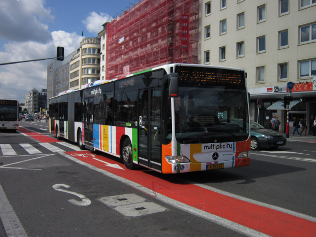 Eine Stadtbus fährt durch eine Straße mit hohen Gebäuden, mit Menschen auf dem Gehweg, Ampeln und Texttafeln, unter einem bewölkten Himmel.