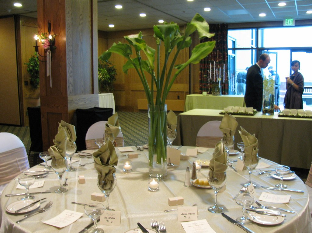 Hotel-Essbereich mit einem weißgedeckten Tisch, der mit Tellern, Besteck und Gläsern sowie einem grünen Tisch im Hintergrund, einem Mann in einem schwarzen Blazer, einem Mädchen neben ihm und einer Tür mit zwei Männern dahinter ausgestattet ist.