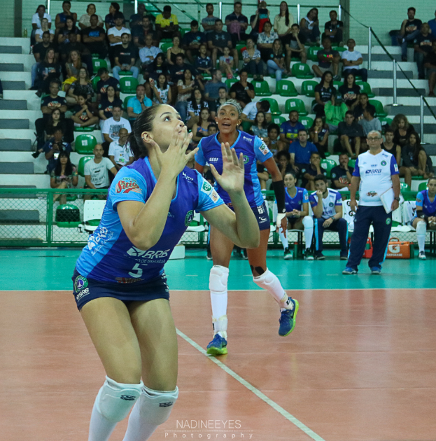 Eine Gruppe von Frauen, die Volleyball auf einem Feld spielen, mit einem Mann auf der rechten Seite, Zuschauern im Hintergrund und Text am unteren Rand des Bildes.