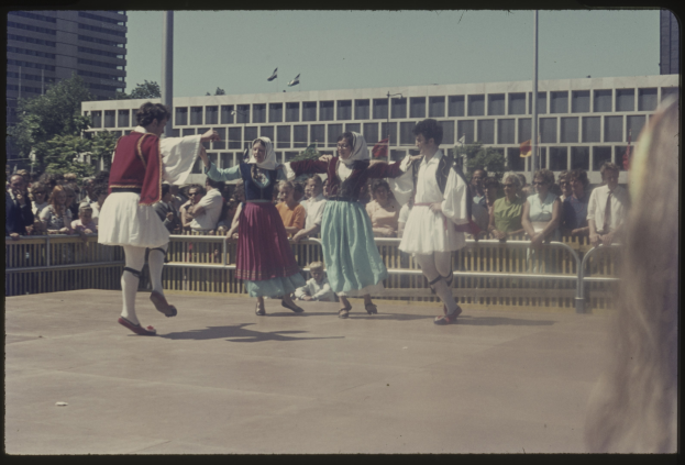 Eine Gruppe von Menschen, die auf einer Bühne tanzen, mit einer Menschenmenge davor, umgeben von Gebäuden, Bäumen, Pfählen, Flaggen und einem klaren blauen Himmel im Hintergrund, alle tragen Schuhe.