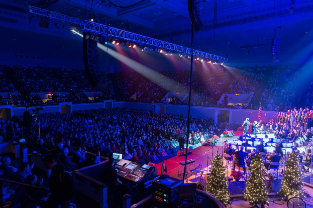 Eine große Menschenmenge in einem geschmückten Auditorium schaut einer Band zu, die auf einer festlichen Bühne mit Weihnachtsbäumen, Lautsprechern und beleuchteter Decke spielt.