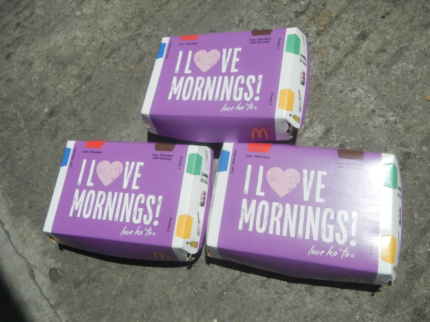 Drei buntfarbene McDonald's 'Ich liebe morgens'-Schachteln mit dem McDonald's-Logo und fetter weißer Schrift auf einem hellblauen Himmel mit weißen Wolken.
