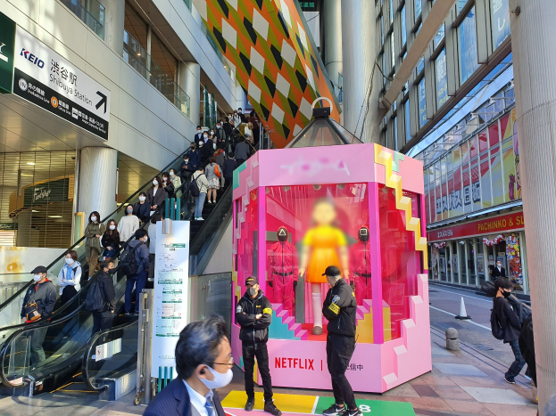 Eine Gruppe von Menschen steht vor einem Gebäude mit Glaswänden, Säulen und Texttafeln; einige tragen Masken, und eine Rolltreppe mit Menschen ist zu sehen. In der Mitte befindet sich ein pinker Kasten, auf dem "Netflix's new Netflix booth in Tokyo" steht.