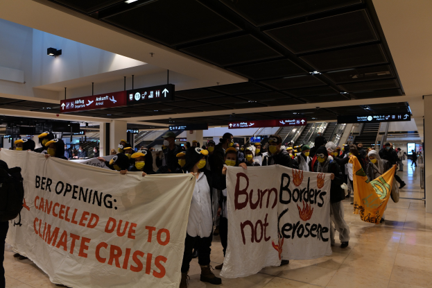 Eine Gruppe von Menschen mit Masken hält Schilder mit der Aufschrift "Verbrenne Grenzen nicht Kerosee" in einem Flughafen mit Rolltreppen, beleuchteter Decke und Texttafeln im Hintergrund.