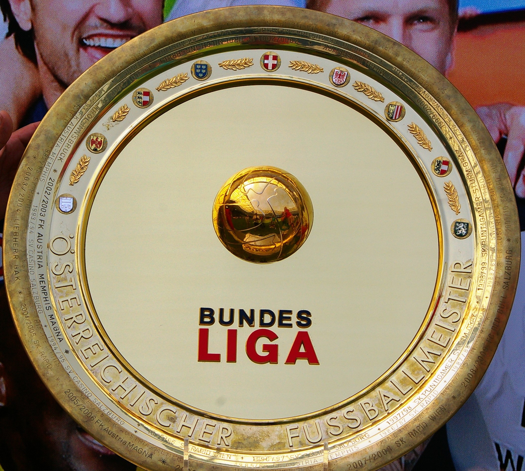 Goldener Schild mit 'Bundesliga'-Text, gehalten von einem Mann mit einem Banner im Hintergrund, das drei Personen zeigt.