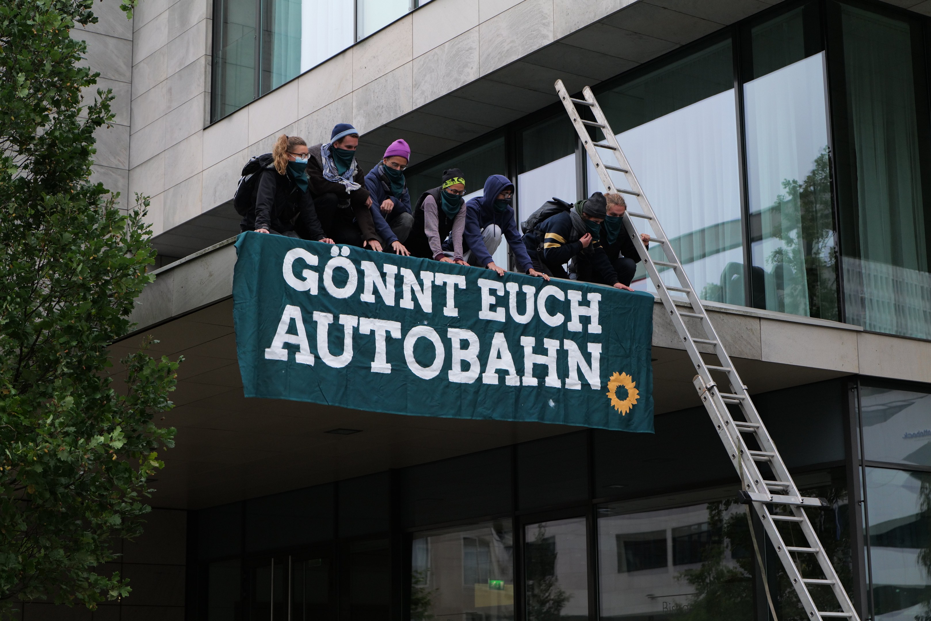 Gruppe von Menschen auf einem Gebäude, die eine Fahne halten, mit einem Baum links und Glasfenstern im Hintergrund, wahrscheinlich protestierend im Namen der deutschen Regierung.