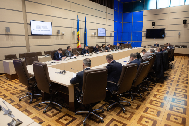 Große Gruppe von Menschen an einem langen Tisch mit Mikrofonen und Papieren während einer Sitzung des Europäischen Parlaments in Brüssel, mit Flaggen, Fernsehgeräten und Deckenleuchten im Hintergrund.