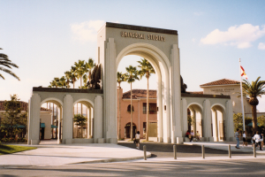 Eingang zum Universal Studios Hollywood mit einem Bogen, einer Statue, Menschen, Bäumen, Gebäuden, einem Fahnenmast und einem bewölkten Himmel.