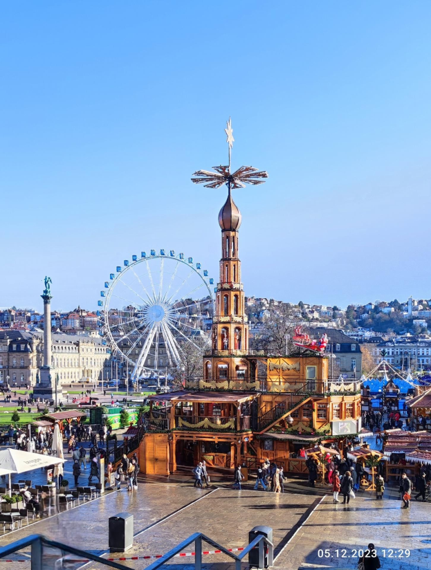 Ein belebter Stadtplatz mit einem zentralen Riesenrad, umgeben von Gebäuden, Bäumen und Menschen unter einem klaren blauen Himmel, mit Marktzelten und Geländern und einem Wasserzeichen unten.