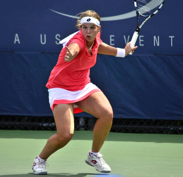 Eine Frau in einem pinken Shirt und weißen Schuhen, die ein Headband trägt, spielt Badminton auf einem Court und hält einen Schläger, während im Hintergrund ein Banner zu sehen ist.