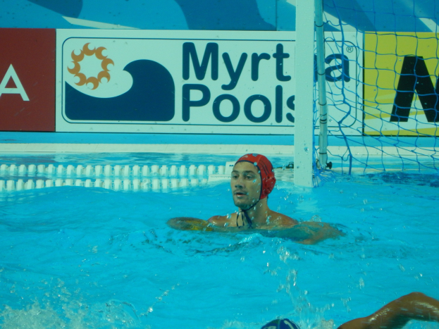 Wasserballspieler in Bewegung in einem Pool, tragend eine Mütze, mit einem Netz auf der rechten Seite und einer Werbetafel im Hintergrund.