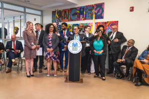 Eine Gruppe von Menschen steht vor einem Podium mit einem Mikrofon und einer Tafel mit der Aufschrift "Sancho-Stadtrat tritt dem Stadtrat bei", mit anderen im Hintergrund sitzend und stehend, Flaggen, einer Tafel, einem Feuerlöscher und Deckenleuchten an der Rückwand.