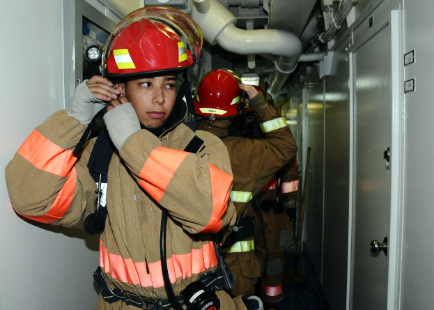 Gruppe von Feuerwehrleuten in Uniform, die zusammen in einem Raum mit einer Tür auf der rechten Seite und einer Wand auf der linken Seite stehen, mit Rohren und Equipment im Hintergrund während eines Trainings.