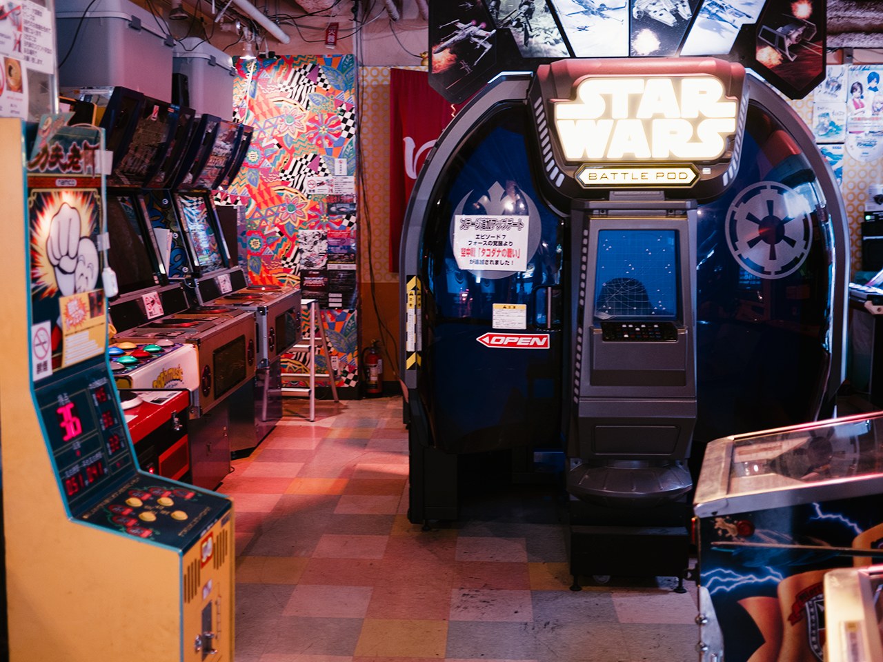 Ein hell erleuchteter Arcade-Raum mit mehreren Maschinen, darunter ein Star Wars Battle Pod, bunten Postern an den Wänden und verstreuten Gegenständen, die eine lebendige Atmosphäre schaffen.