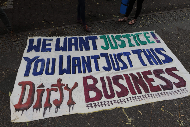 Banner auf der Straße mit Text "Wir wollen Gerechtigkeit, du willst nur diesen schmutzigen Deal" und zwei Personen oben drauf, eine hält ein blaues Objekt.