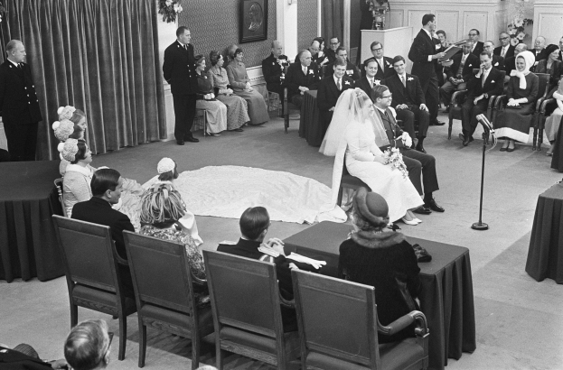 Schwarzes und weißes Foto einer Hochzeitszeremonie mit sitzenden und stehenden Menschen, einem Mann mit Mikrofon, einer Frau mit Blumen, einem Podium mit Mikrofon, Blumenschmuck, einem Vorhang, einem gerahmten Foto und einer Tür im Hintergrund.