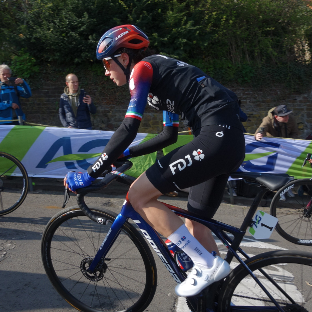 Professionelle Radsportlerin Katie Archibald mit Helm, Schutzbrille, Handschuhen und Schuhen fährt auf einem Fahrrad eine Straße entlang neben einer Menschenmenge, mit einem Banner, einer Wand, Bäumen und einem Gebäude im Hintergrund.