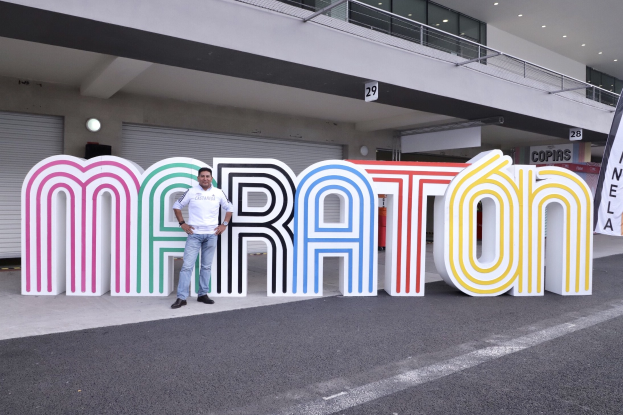 Ein Mann steht am Start eines Marathons vor einem großen "Marathon"-Schild, mit einem Banner rechts daneben und einem Gebäude mit Lichtern, Geländern und Läden im Hintergrund.
