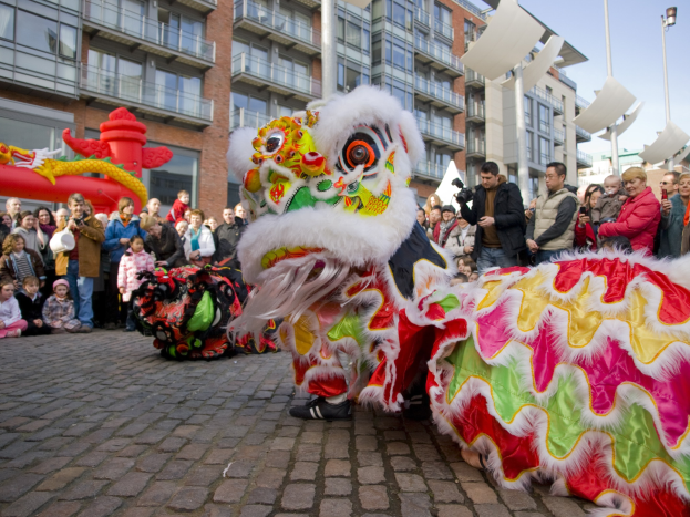 Ein lebendiges chinesisches Neujahrsfest in Amsterdam mit einem Löwen tanzen vor einem Publikum, einige fotografieren das Ereignis, vor einer Kulisse aus Gebäuden, Laternenmasten und einem klaren blauen Himmel.