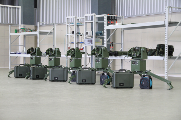 Gruppe von Milit├Ąrrobotern auf einem Boden mit Koffern und Gegenst├Ąnden auf Gestellen, in der N├Ąhe einer Wand und eines Gel├Ąnders als Teil eines Verteidigungsystems.