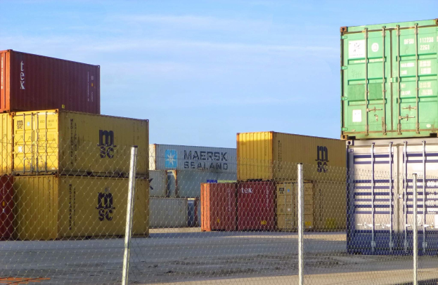 Container auf einem Stapel mit Maschendrahtzaun im Vordergrund und einer Straße darunter, unter einem bewölkten Himmel.