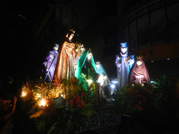 Eine Gruppe von Menschen auf einem schwimmenden Ger├Ąt bei einer nächtlichen Parade, umgeben von Pflanzen, mit einem Stadtgebäude im Hintergrund.