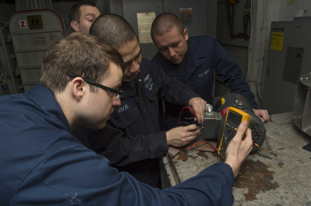Vier Männer in einem Raum, die an einer Maschine arbeiten, mit einem Tisch voller Objekte rechts und Hintergrundmaschinen und Papieren an der Wand.