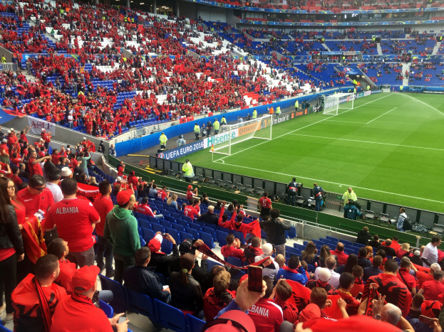 Eine große Menschenmenge in einem Stadion schaut einem Fuballspiel zu, mit Sitzplätzen und Stehplätzen, Bannern, einem Torpfosten und Deckenbeleuchtung.