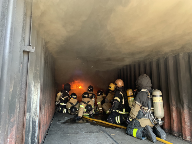 Feuerwehrleute in Schutzausrüstung mit Sauerstoffflaschen arbeiten in einem raucherfüllten Tunnel, einer hält einen Schlauch, mit sichtbaren Flammen im Hintergrund und einer Wand links.