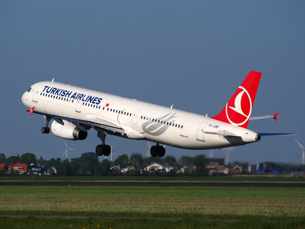 Turkish Airlines Airbus A330-300 startet von Frankfurt Airport, Gras am Boden, Gebäude und Bäume im Hintergrund, Windmühlen in der Ferne, unter einem klaren blauen Himmel.