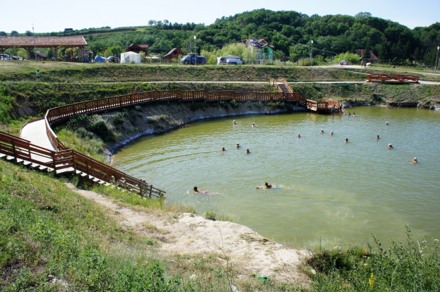 Menschen schwimmen in einem Gewässer umgeben von Grün mit einer Brücke, Treppen, Hütten, Fahrzeugen, Pfählen und einem klaren Himmel im Hintergrund.