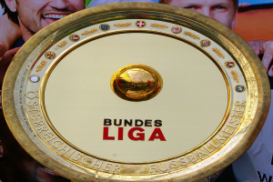 Goldener Schild mit 'Bundesliga'-Text, gehalten von einem Mann, mit einem Banner im Hintergrund mit drei Personen.