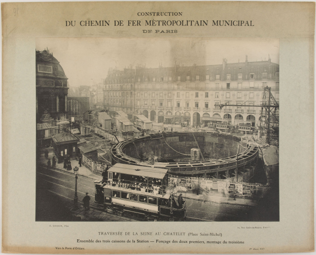 Schwarz-weiß-Foto einer Pariser Stadtstraße im Bau mit einem Zug auf den Schienen, umgeben von Gebäuden, Strommasten und Passanten, mit der Aufschrift "Construction du Chemin de Fer Metropolitain Municipal de Paris" oben.