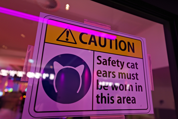 Hellgelbes Warnschild in einem Ladenschaufenster mit der Aufschrift "Vorsicht: Sicherheits-Katzenohren müssen in diesem Bereich getragen werden." vor einem unscharfen Hintergrund.